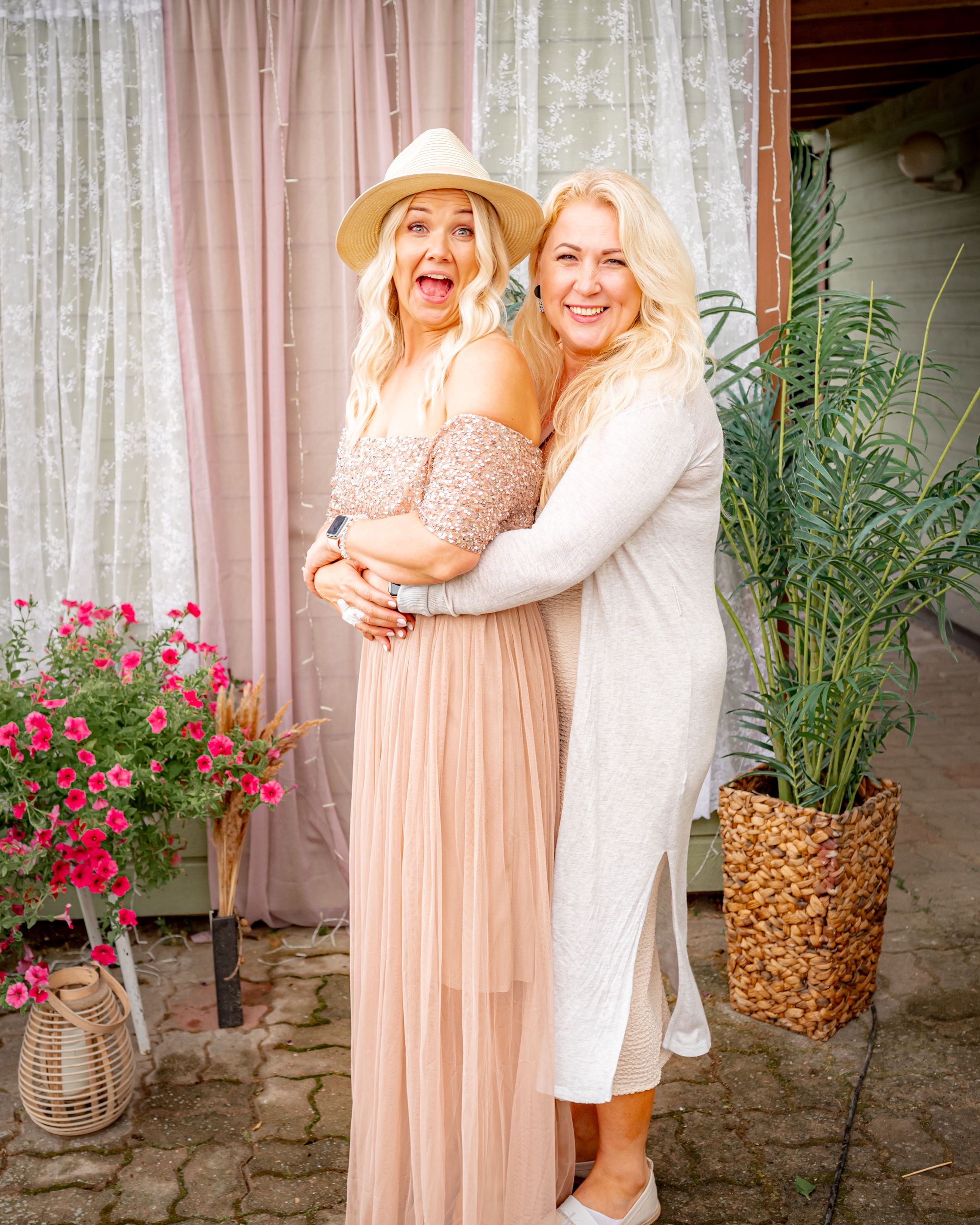Two friends embrace and laugh together in summer outfits against a curtained backdrop with pink flowers and greenery.