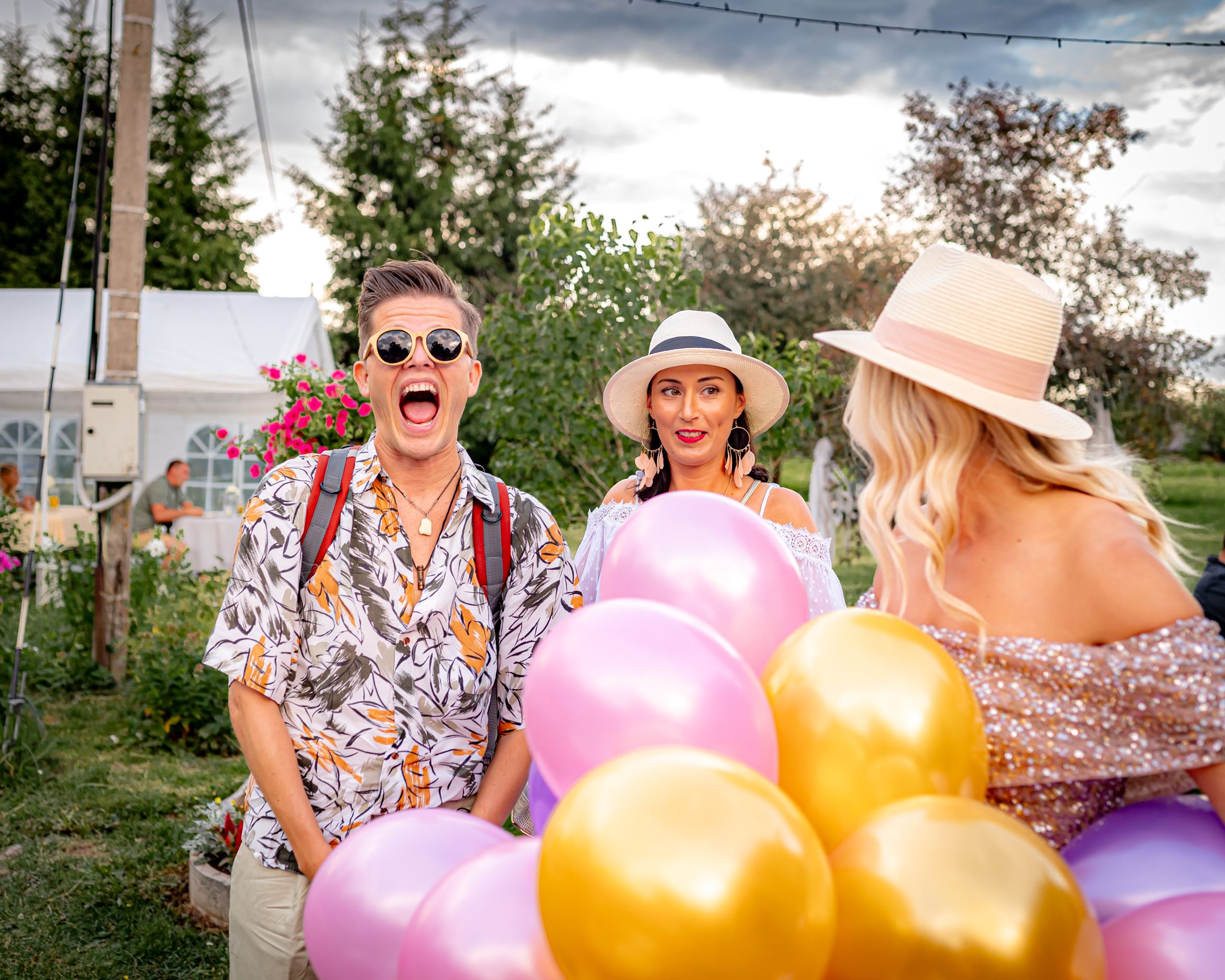 Party guests with colorful balloons celebrate outdoors under string lights at dusk.