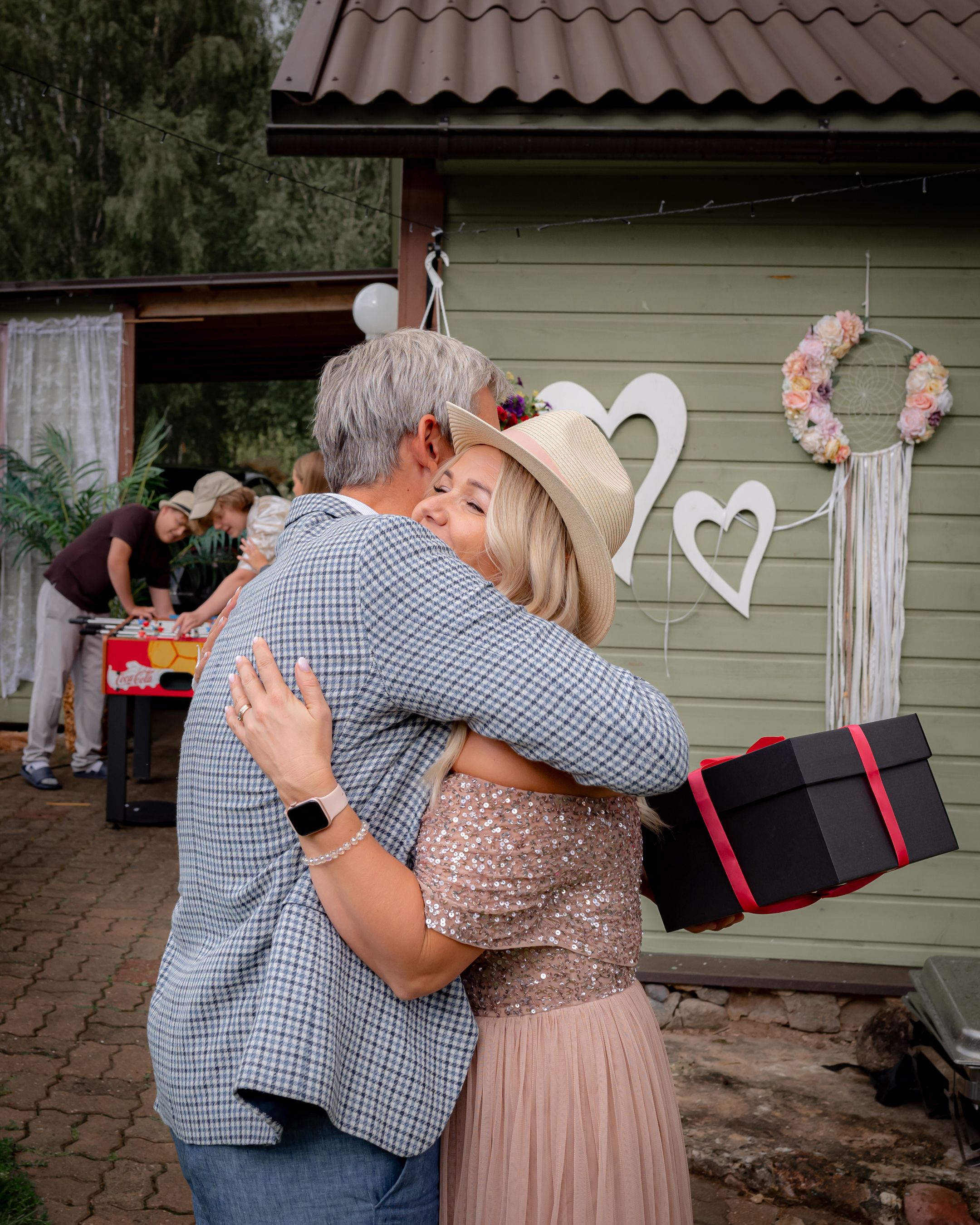 A heartfelt embrace between guests at an outdoor party with heart decorations on the wall.