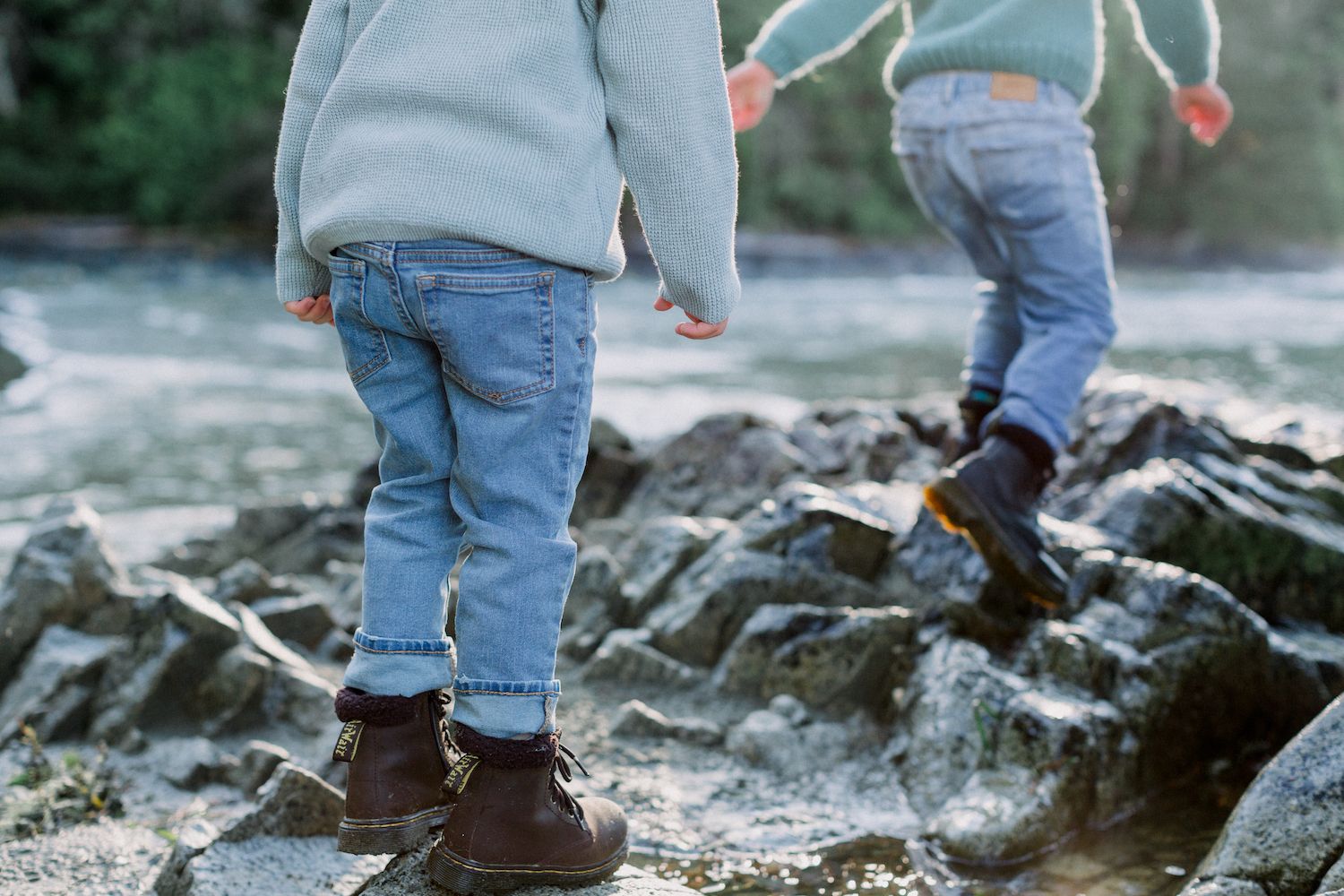Two young children in jeans and boots walking on rocky ground near water.