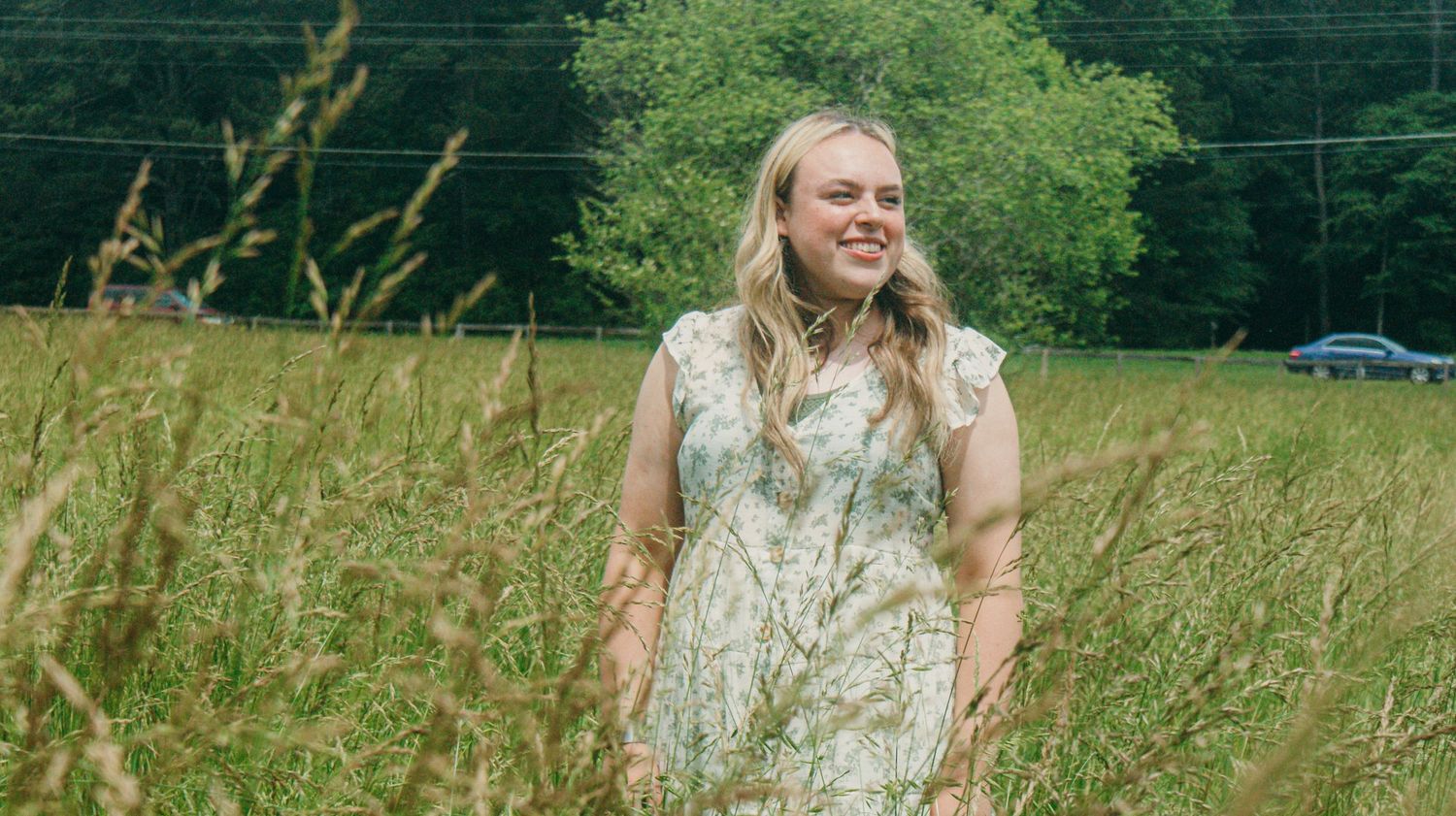 Person in a light colored dress stands among tall wild grasses in a sunny meadow.