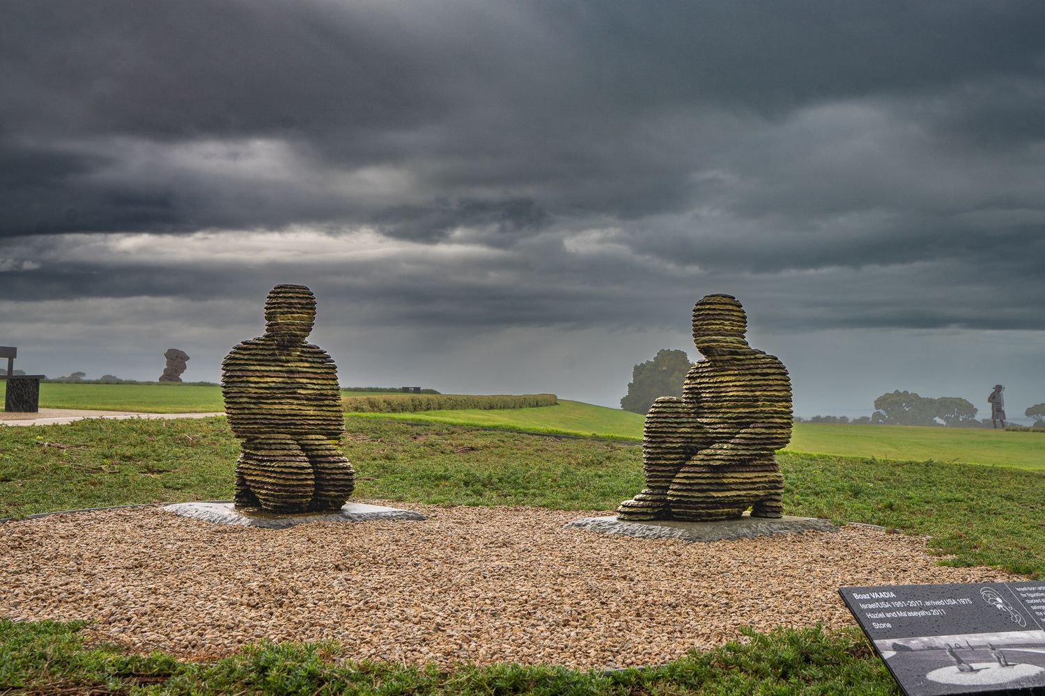 Two stone sculptured human figures kneeling on gravel under dramatic stormy skies in open landscape.