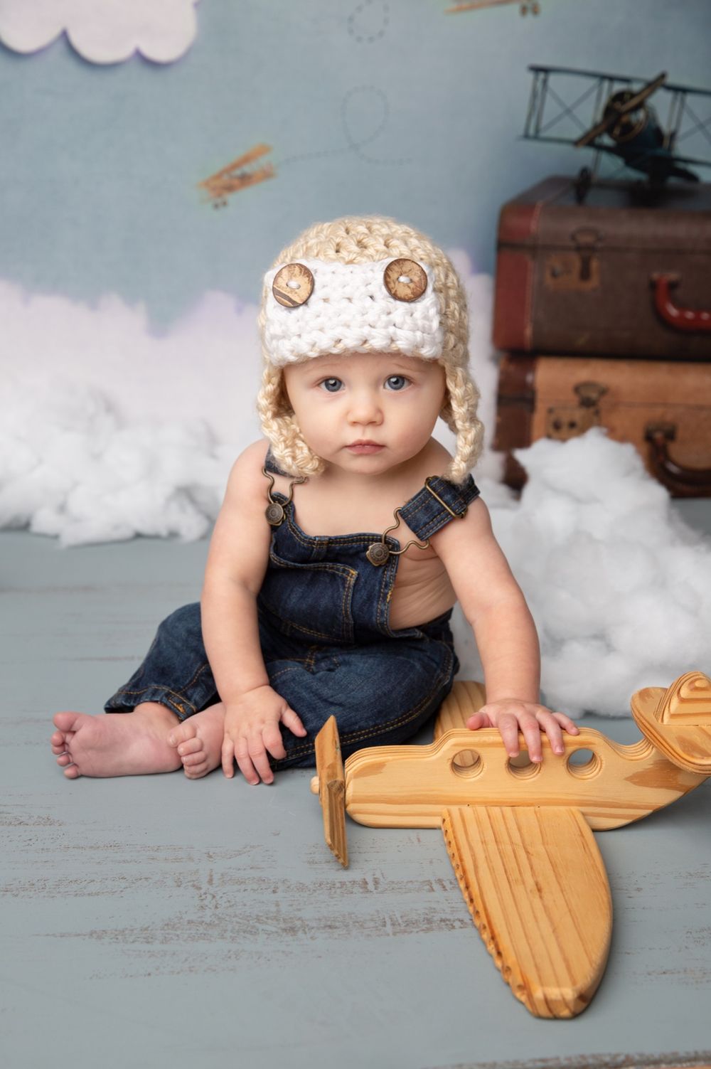 Baby in denim overalls and knit aviator hat sits on floor with wooden toy airplane in vintage aviation themed setting.