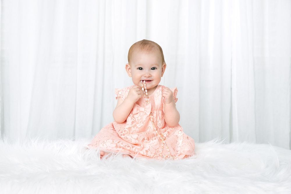 Baby wearing peachy lace dress sits on white fluffy rug against bright background.