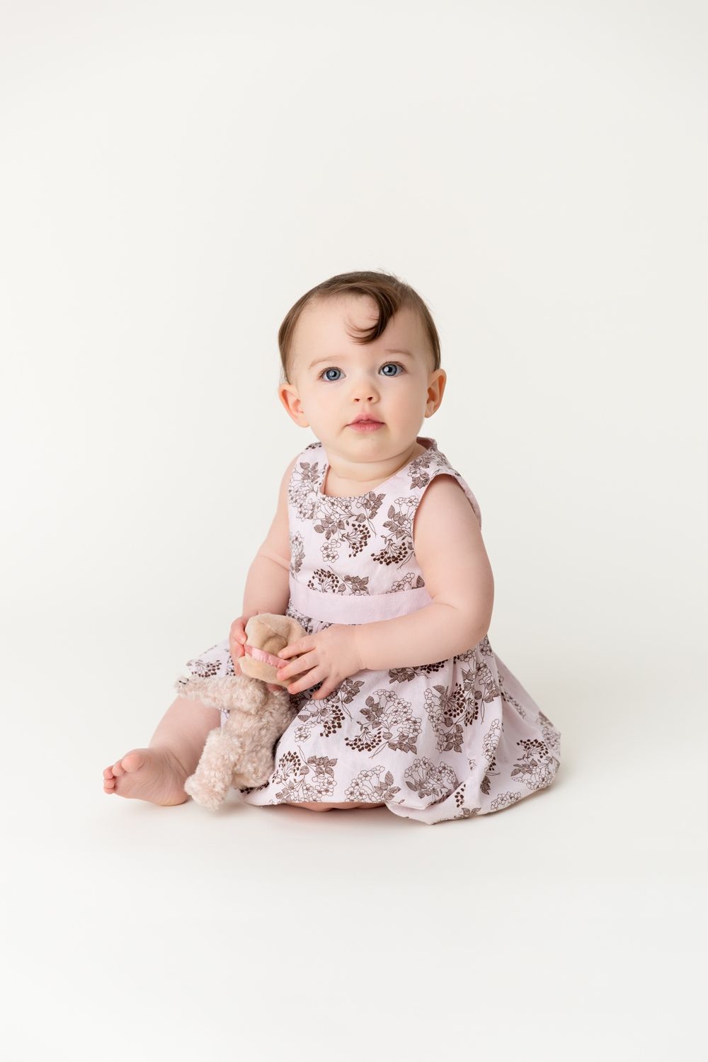 Baby in floral print dress sits with stuffed toy against clean white backdrop.