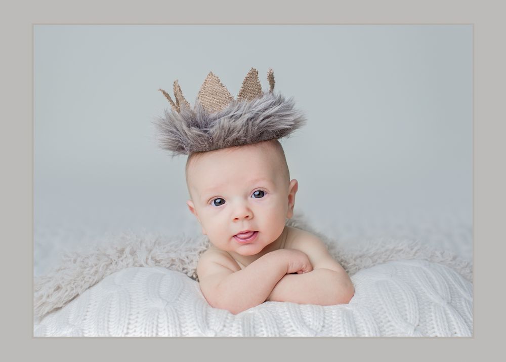 Baby wearing feathered crown lies on white bedding in dreamy portrait setting.