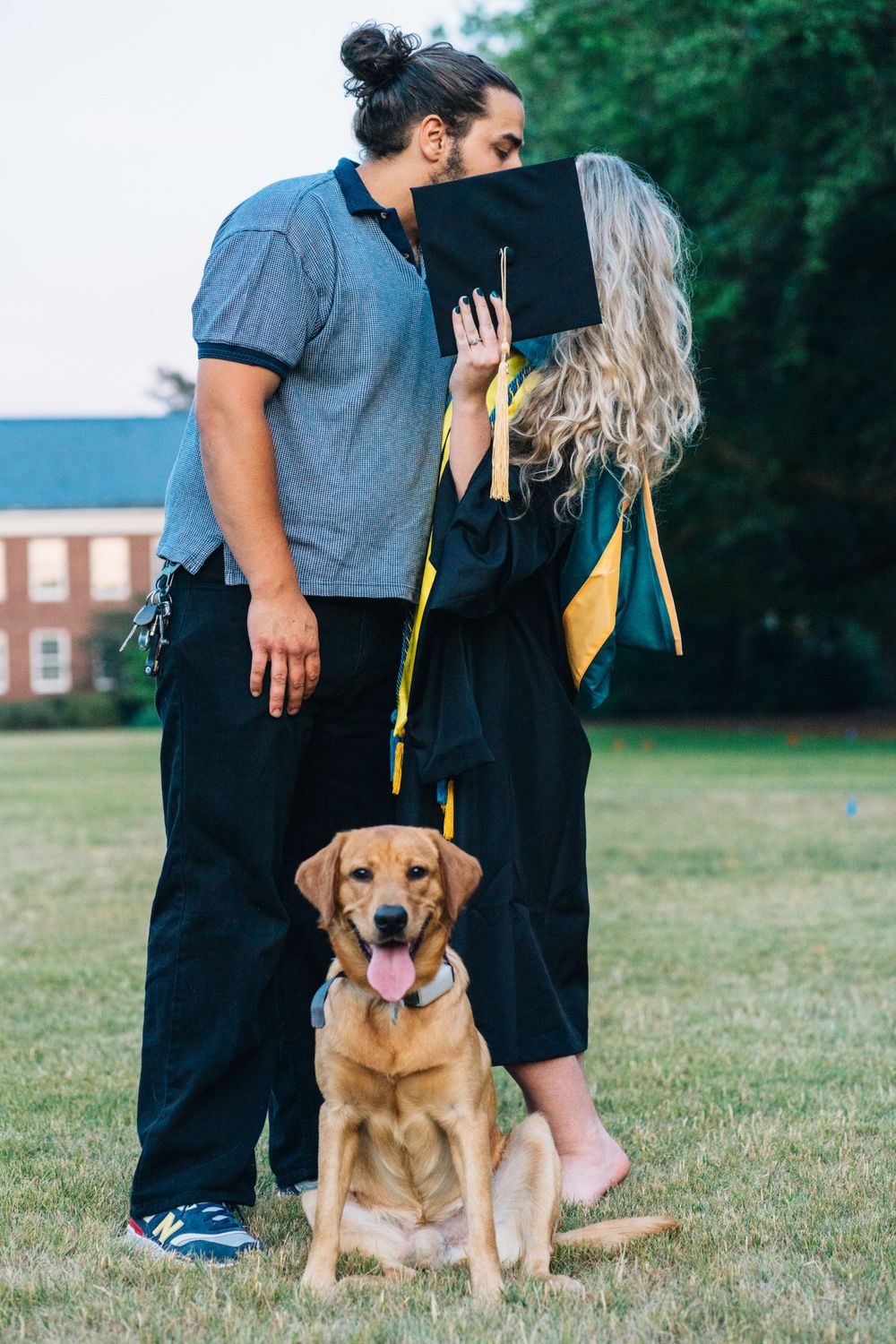 A masters graduate hiding a kiss by her husband with a dog for graduation photos wearing a black dress with gold stole