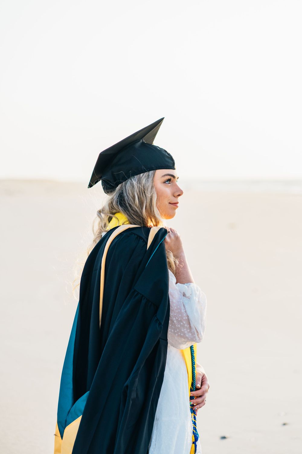 A masters graduate student wearing cap holding gown over shoulder looks into the distance on the beach light and airy