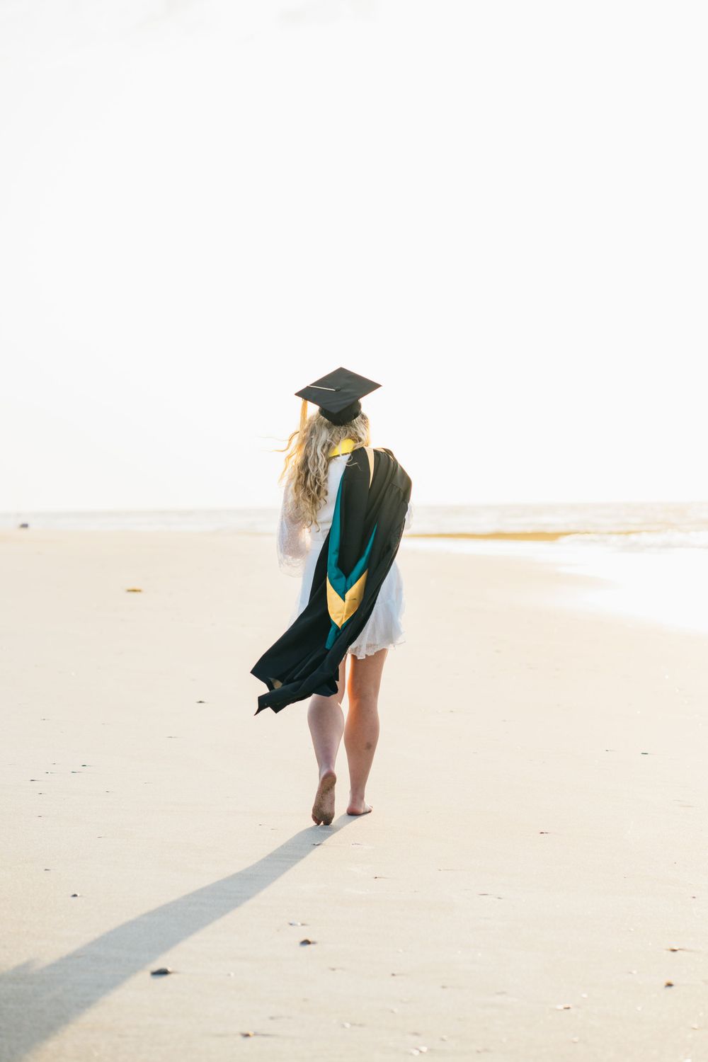 A masters graduate walks on the beach with her cap and gown and white dress, barefoot, light and airy
