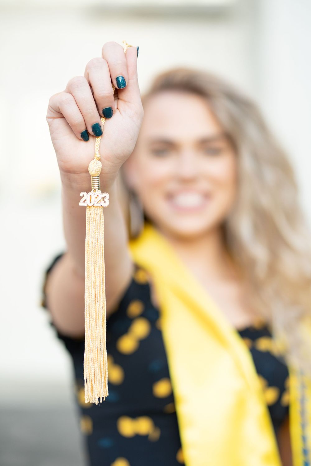 A masters graduate holding her gold tassel in focus in front of an academic building wearing her stoles and cords