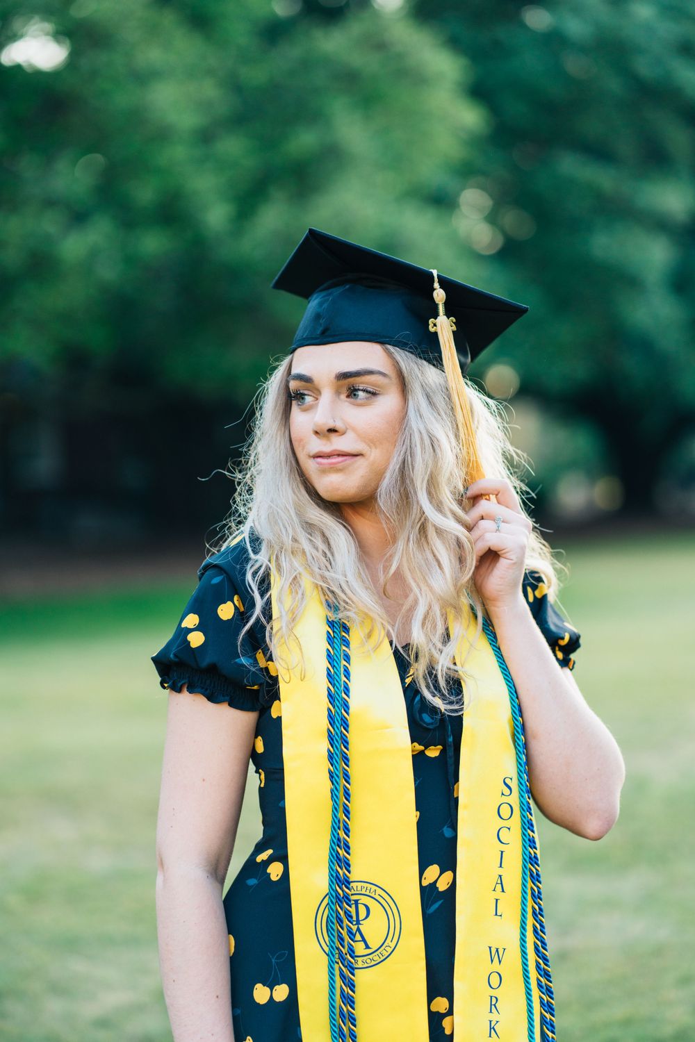 A masters graduate plays with her tassel and cap while wearing stole and cords and dress for graduation photos