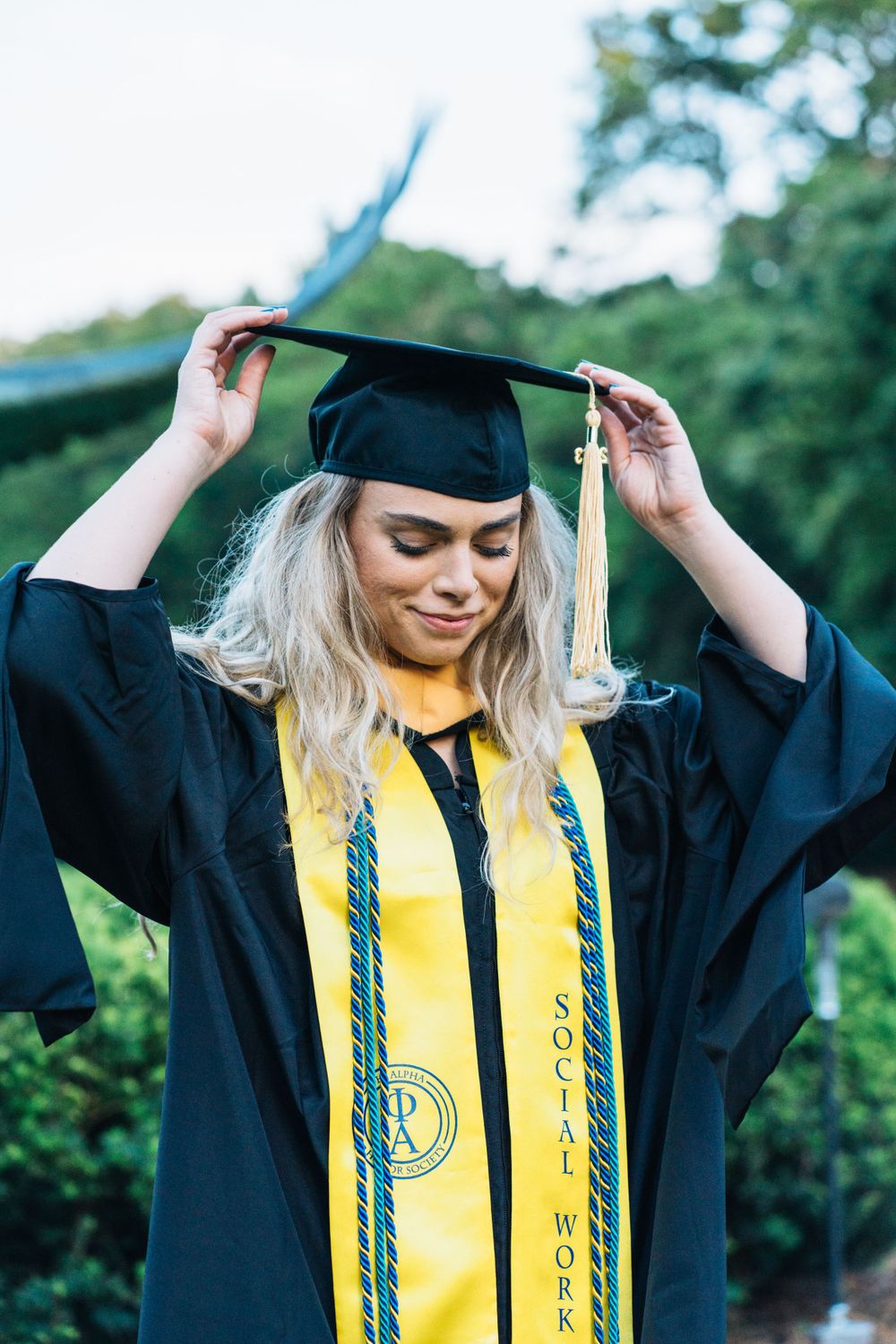 A masters graduate student touching her cap with both hands and looking down in a black dress with full regalia