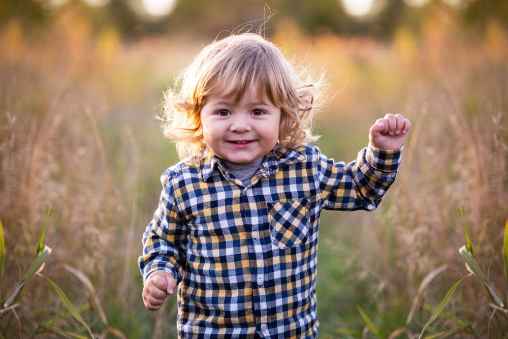 Happy toddler boy with curly hair in a plaid shirt running through a golden field smiling.