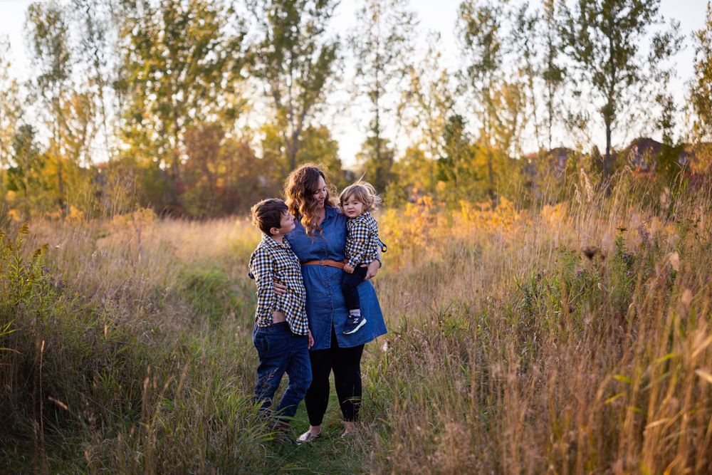 Mother holding young child while older child hugs her in a sunlit autumn field at golden hour.