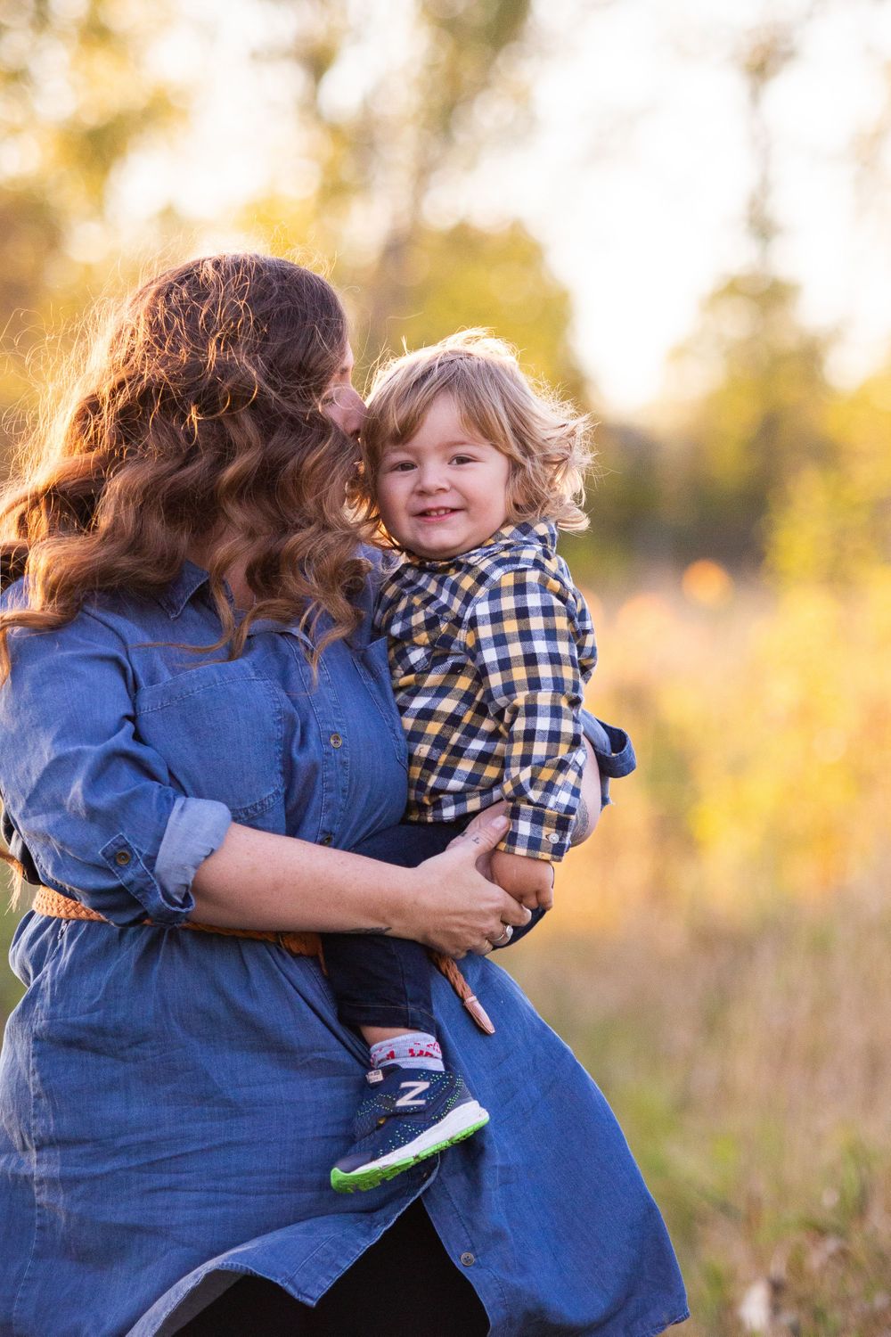 A mother kisses her smiling toddler boy while holding him in a sunny autumn field.