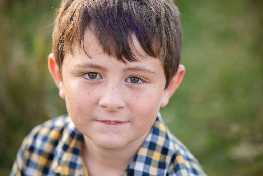 A young freckled boy in a plaid shirt smiles softly in an outdoor portrait.