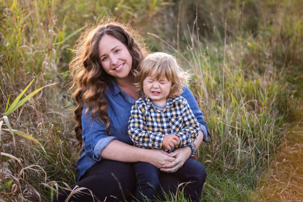 A smiling mother sits in tall autumn grass holding her happy toddler on her lap.