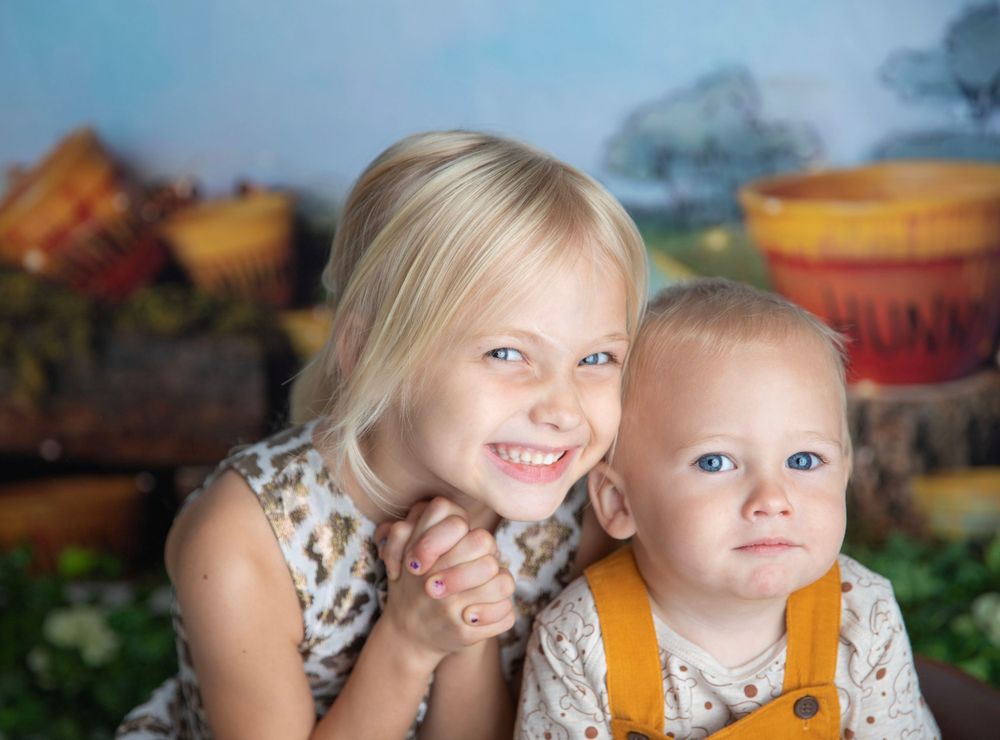 Smiling blonde girl hugging a baby boy in mustard overalls against a colorful storybook backdrop.