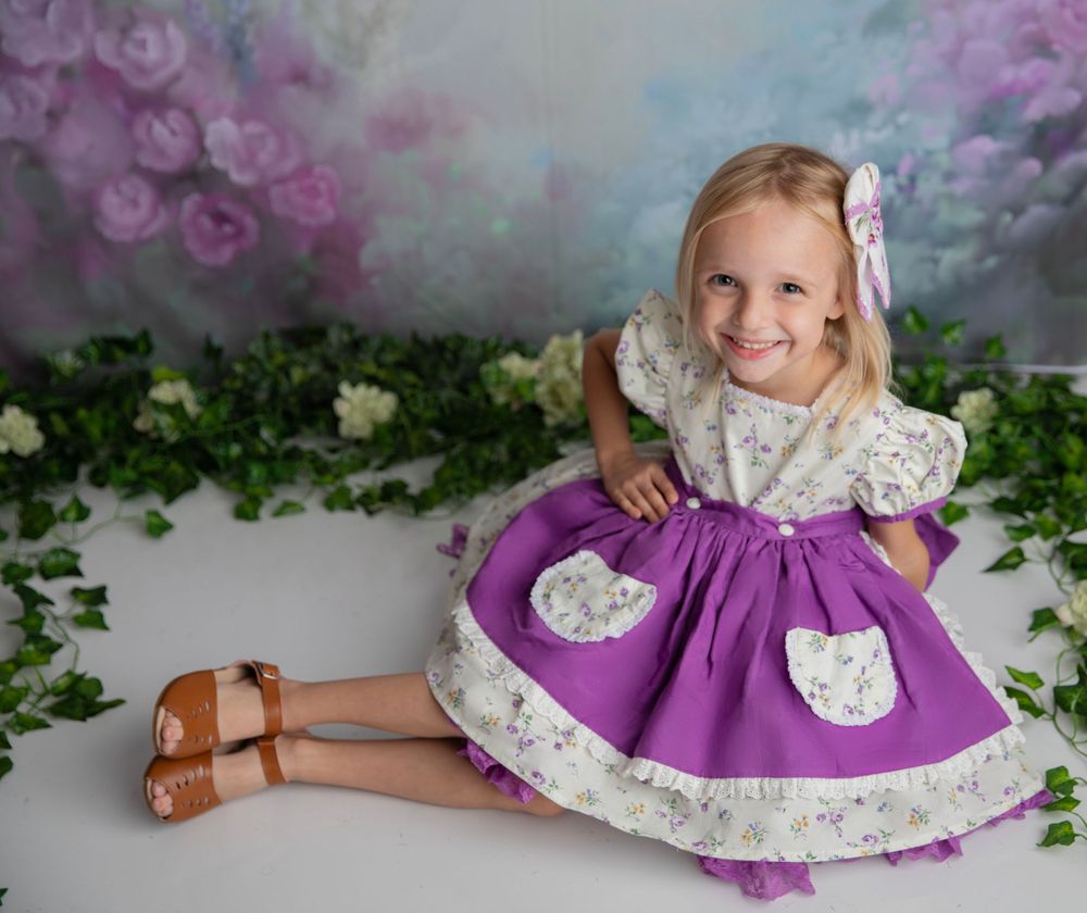 Smiling blonde girl in a purple and white floral dress sitting among green ivy on a floral backdrop.