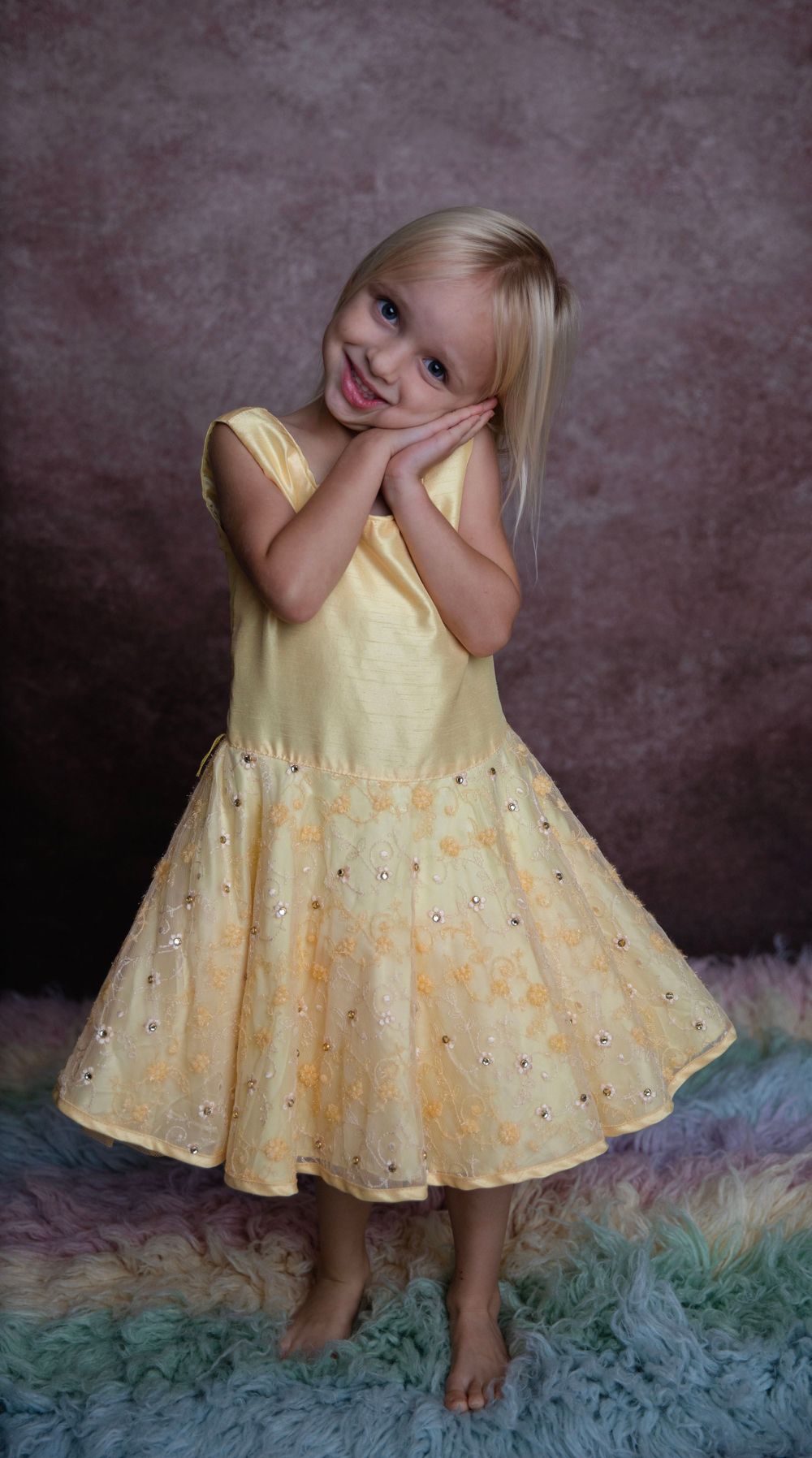 Smiling blonde girl in a yellow sequined dress posing with hands on cheeks on a rainbow fur rug.