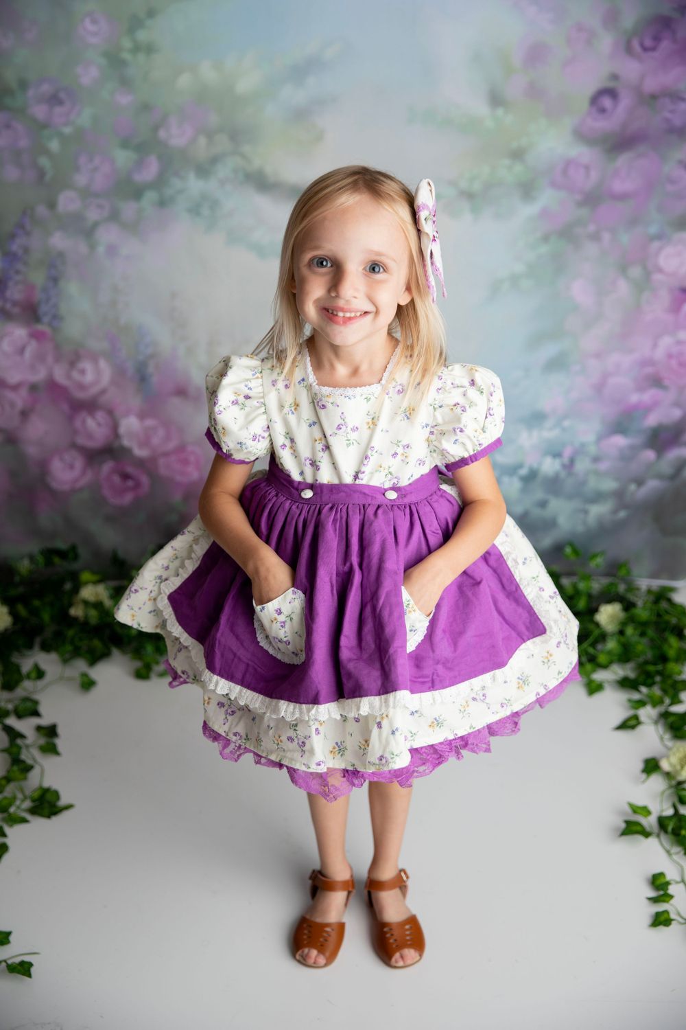 Smiling blonde girl in a purple and white floral dress with pockets standing before a floral backdrop.