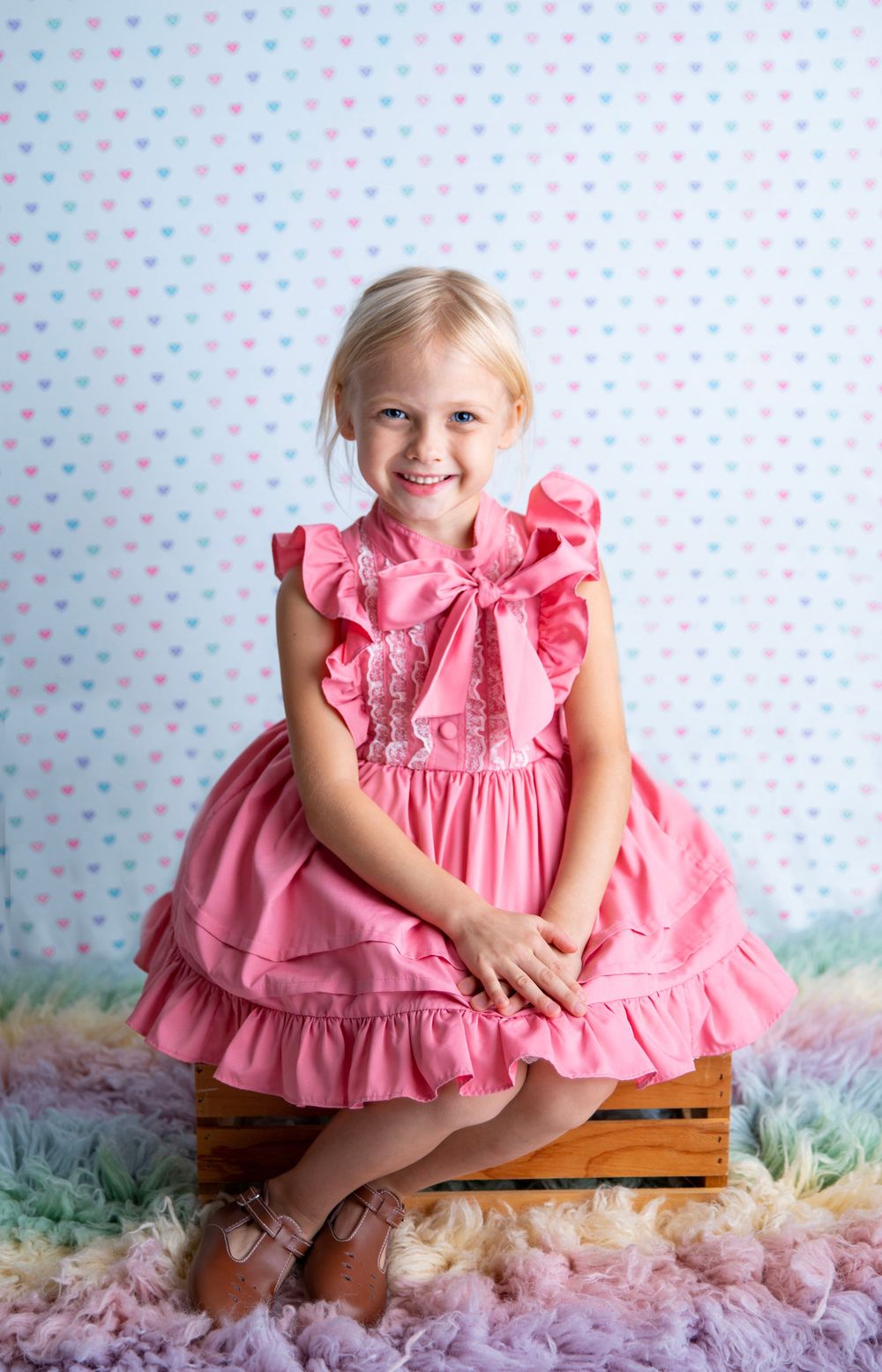 Happy blonde girl in a pink ruffled dress with a bow sitting on a wooden crate over a rainbow fur rug.