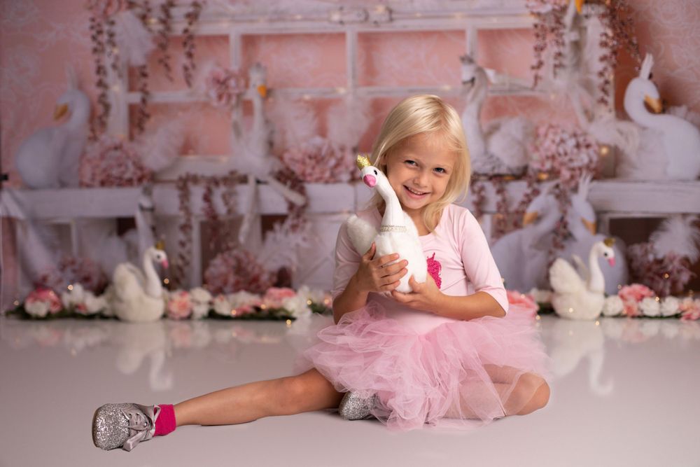 A cheerful little girl in a pink tutu holds a white swan plush toy in a whimsical studio setting.