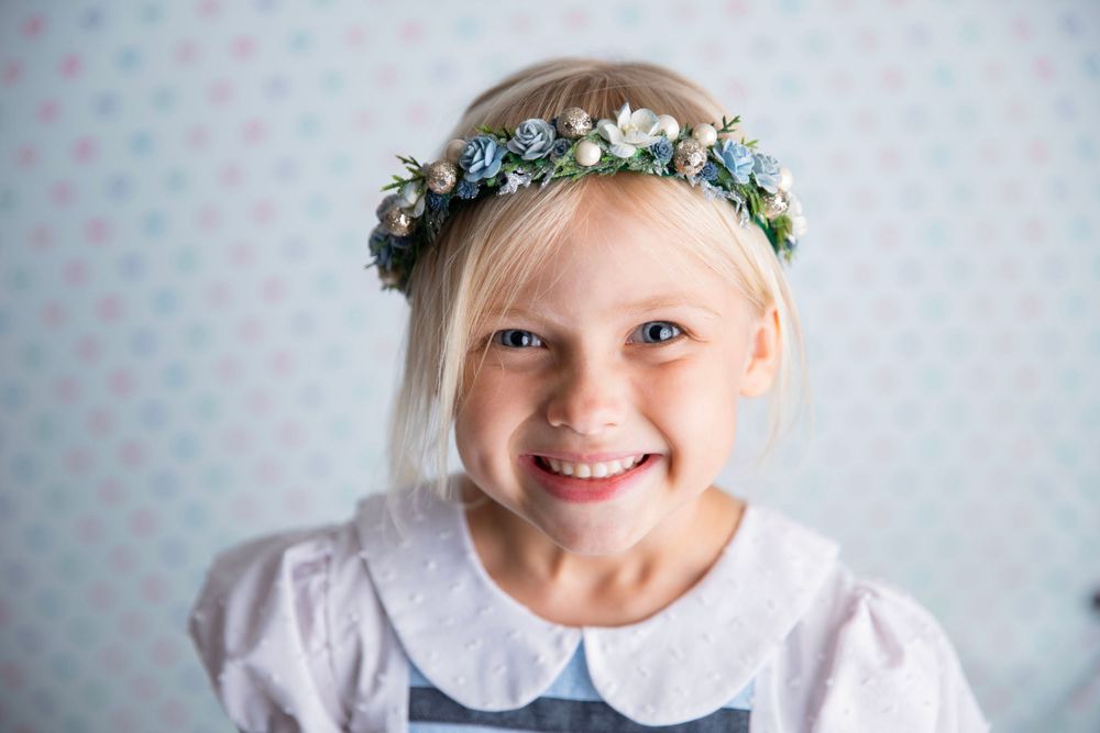 Cheerful blonde girl wearing a floral crown of blue and white flowers smiling at the camera.