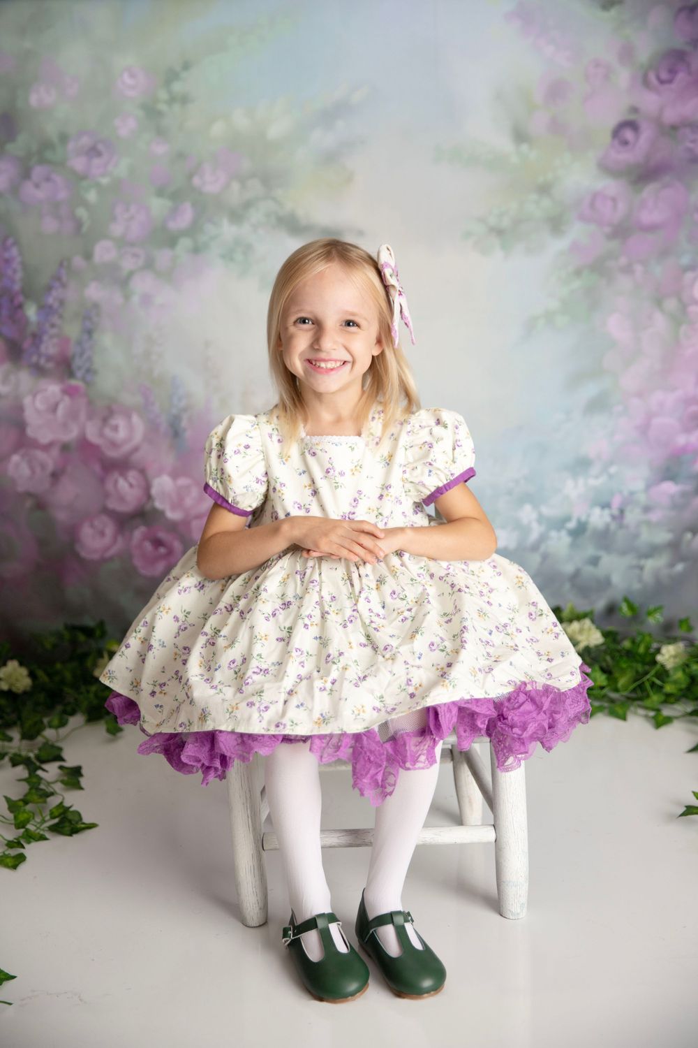 Smiling blonde girl in a floral purple and white dress seated on a stool before a floral painted backdrop.