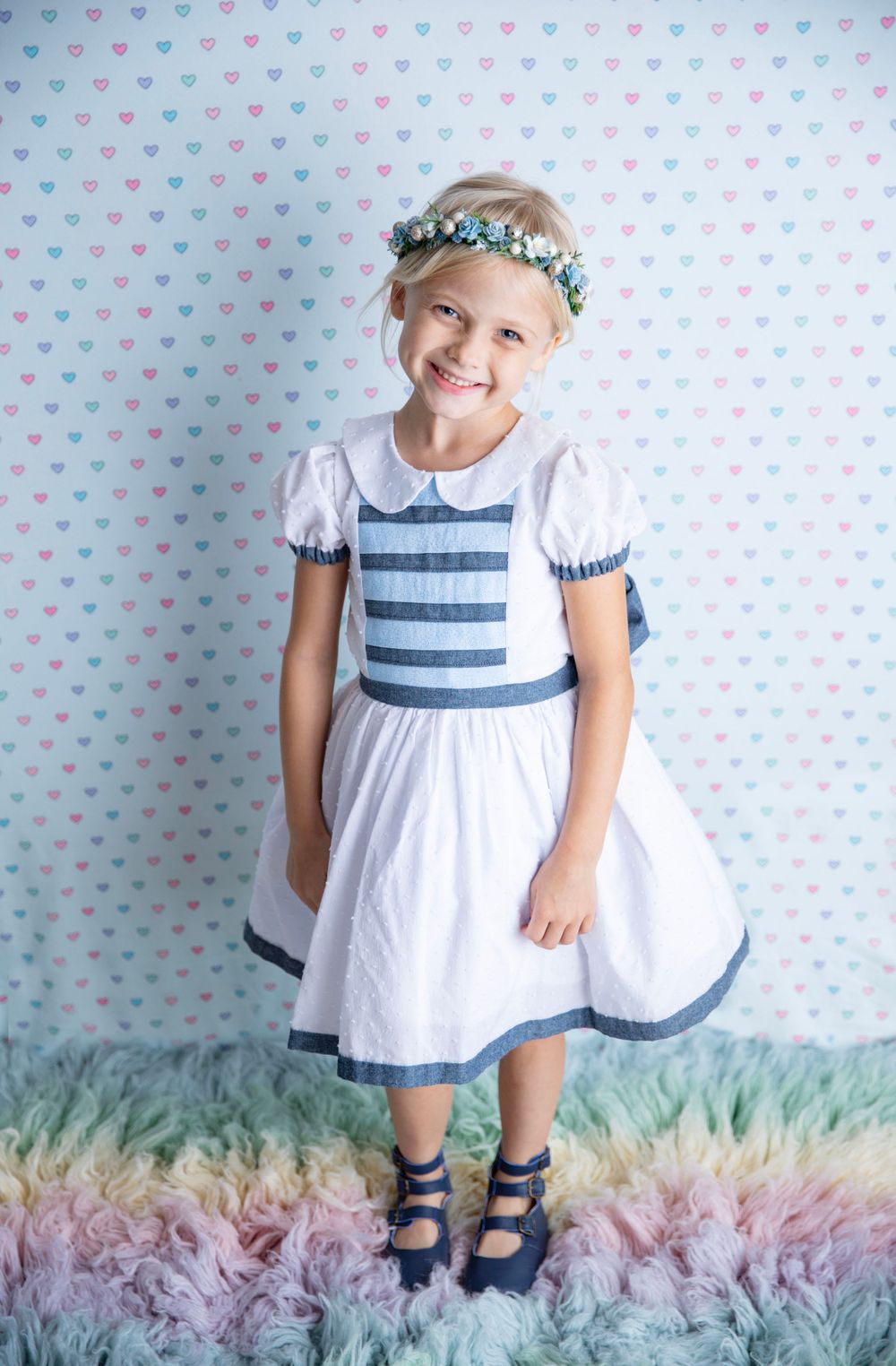 Blonde girl in a white and blue striped dress with a floral crown standing on a rainbow fur rug.