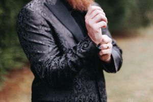 Groom in black floral tuxedo jacket adjusting cufflinks outdoors before wedding.