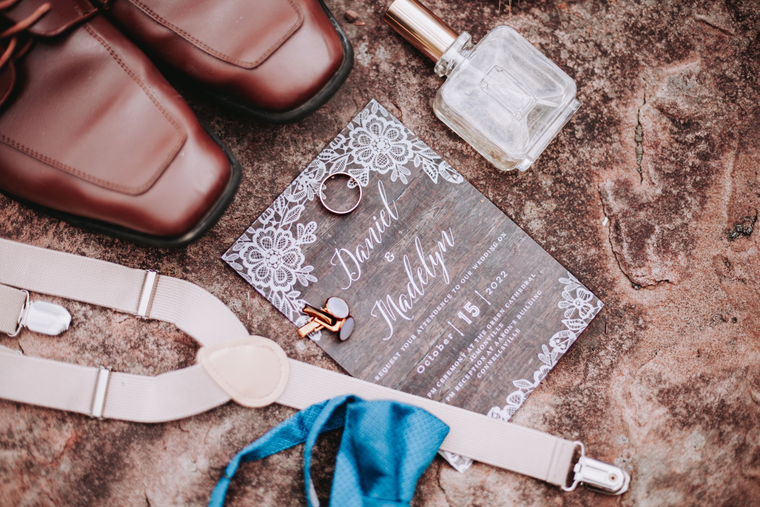 Flat lay of wedding invitation, rings, cufflinks, suspenders, shoes, and cologne on sandy surface.