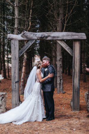 Bride and groom sharing first kiss under a rustic wooden arch in a pine forest ceremony.