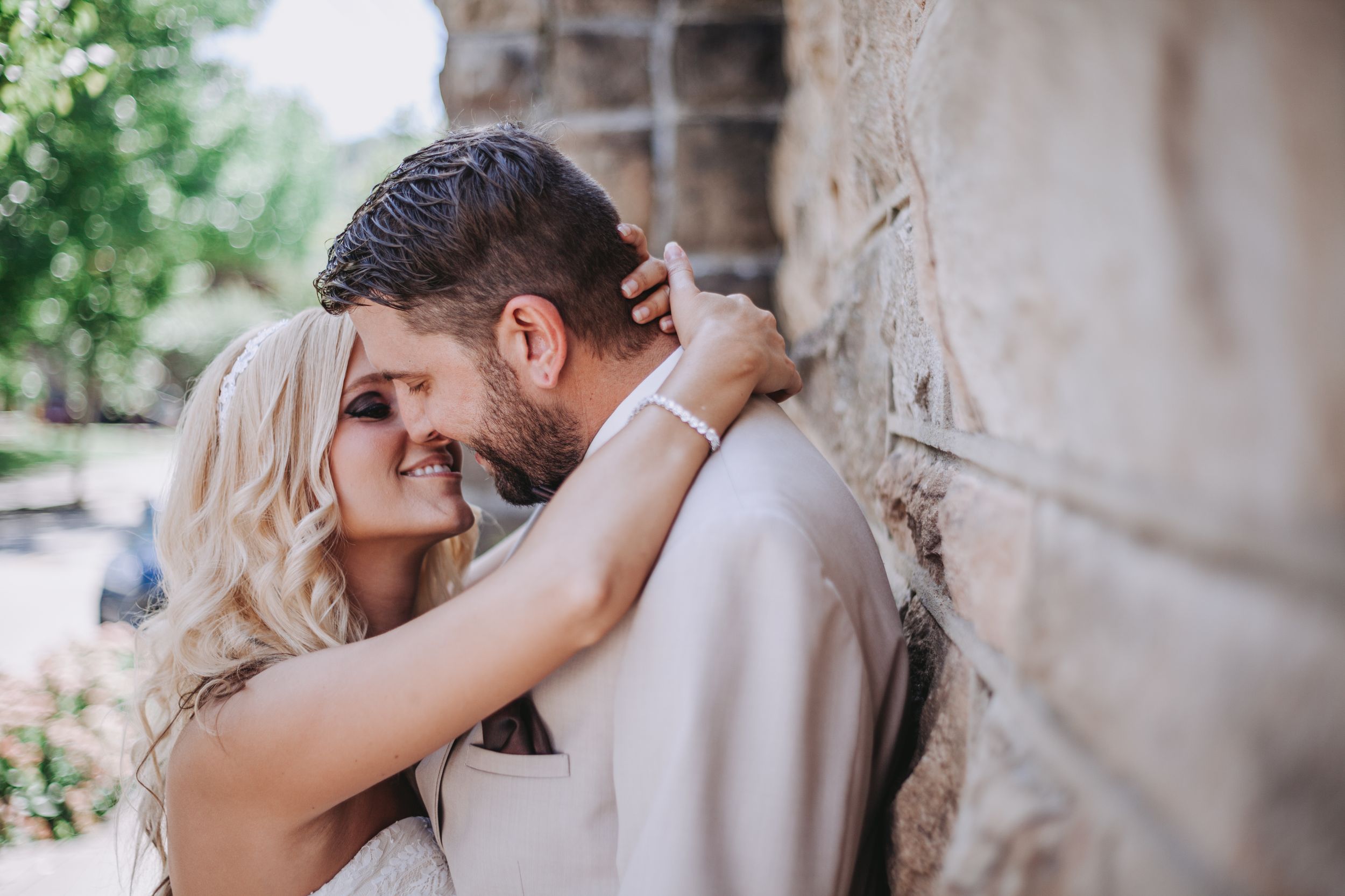 Smiling blonde bride leaning against groom near stone wall in sunny outdoor setting.