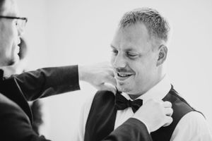 Groomsman adjusts the bow tie of a smiling groom in a black and white wedding preparation photo.