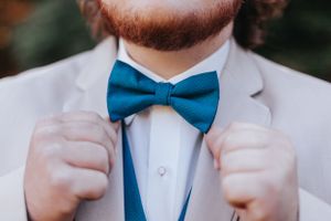 Groom in light gray suit adjusts teal blue bow tie with both hands on wedding day.
