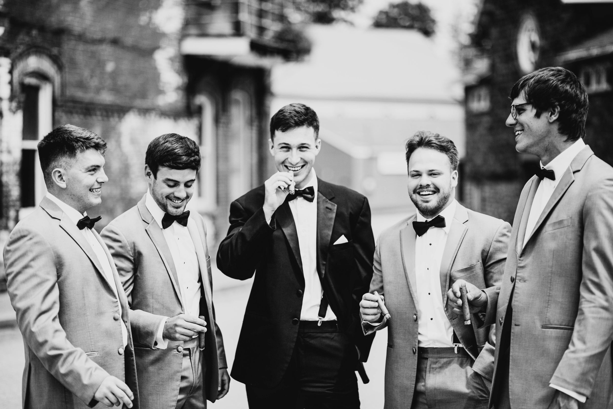 Groom and groomsmen laughing together in suits and bow ties at wedding.