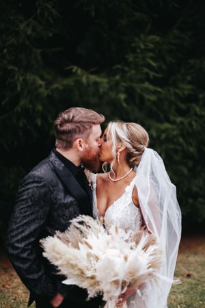 Bride and groom sharing a romantic kiss outdoors with a pampas grass bouquet.