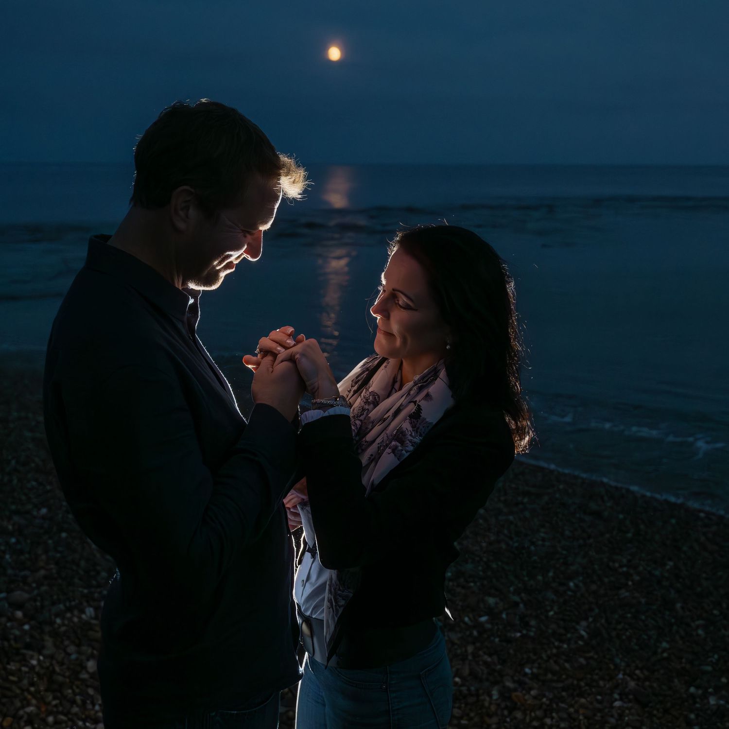 Couple holds hands in romantic moonlit moment on beach with ocean backdrop.