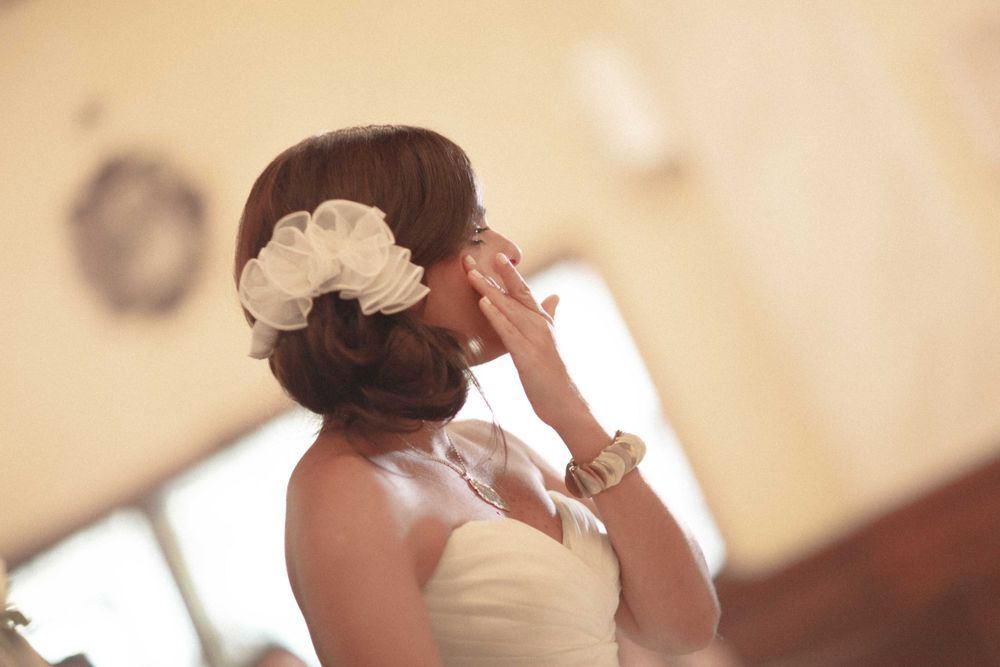 A bride in a strapless wedding dress touches her face while wearing a white floral hair accessory and bracelet.