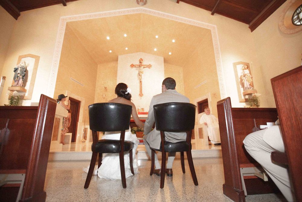 Two people sit in chairs facing a crucifix at the altar of a small chapel with wooden beams and religious artwork.