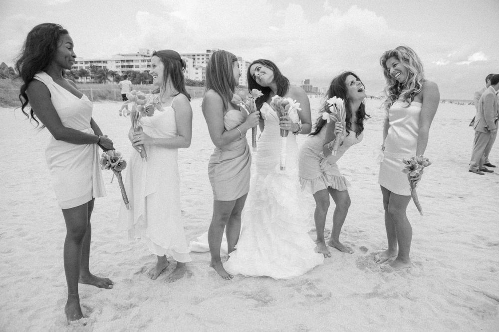 A group of women in light-colored dresses laugh and celebrate together on a beach during a sunny day.