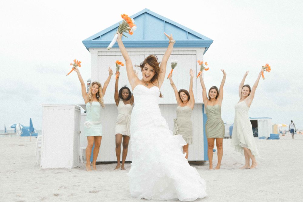 A group in white dresses jumps joyfully on a sunny beach with a blue beach house in the background.