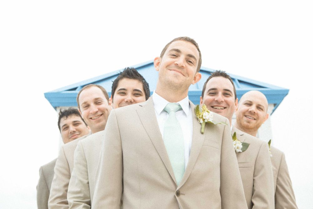 Groomsmen in beige suits and light blue ties smile together under blue sky on wedding day.