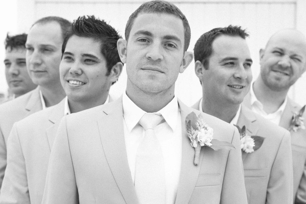 A group of groomsmen in light colored suits stand together and smile at an outdoor wedding celebration.