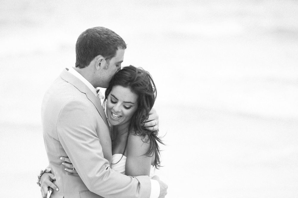 A romantic couple in white clothing embrace together in soft sunlight on the beach.