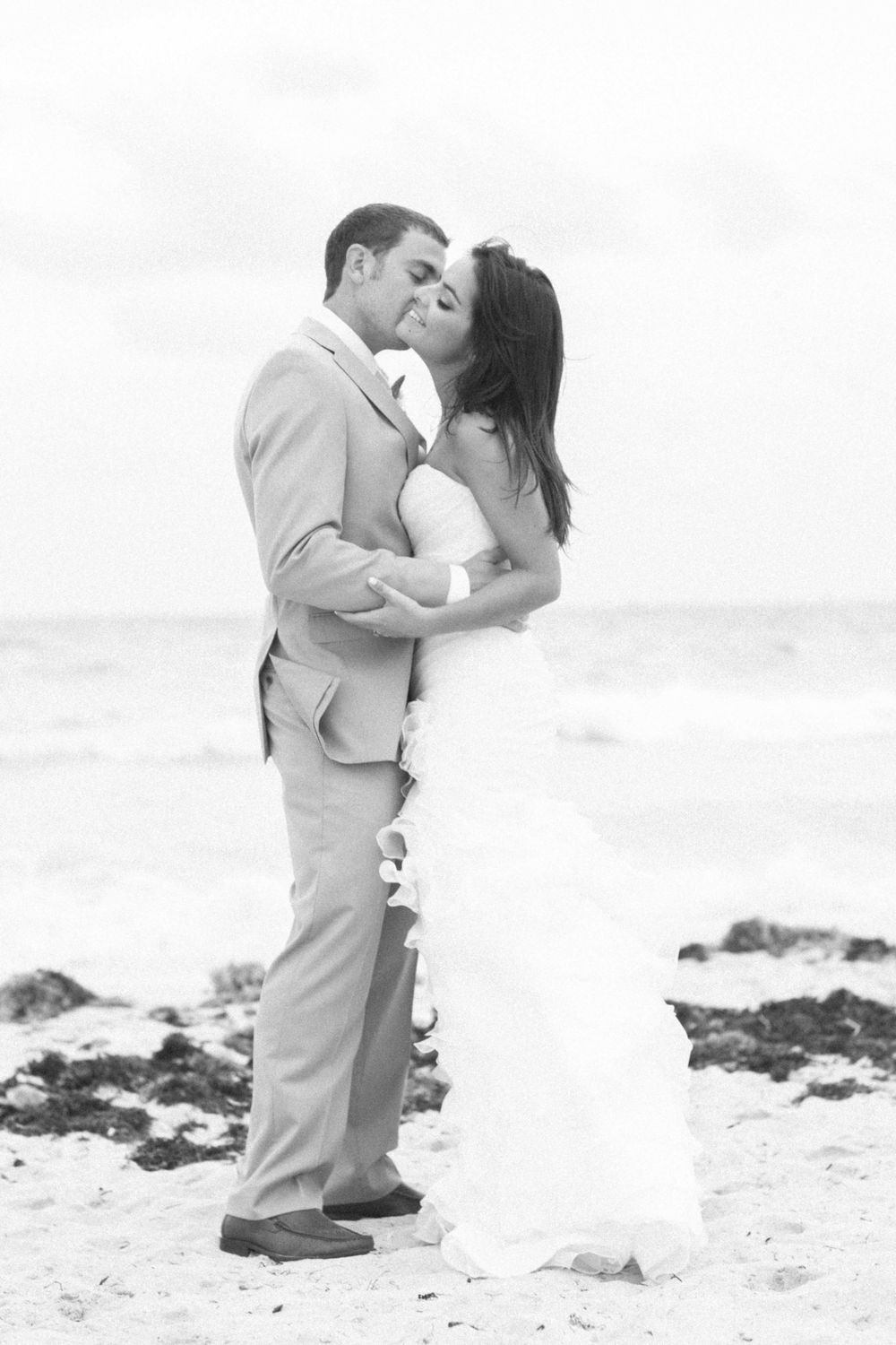 A romantic black and white beach wedding photo of newlyweds kissing on the sand with ocean waves in the background.