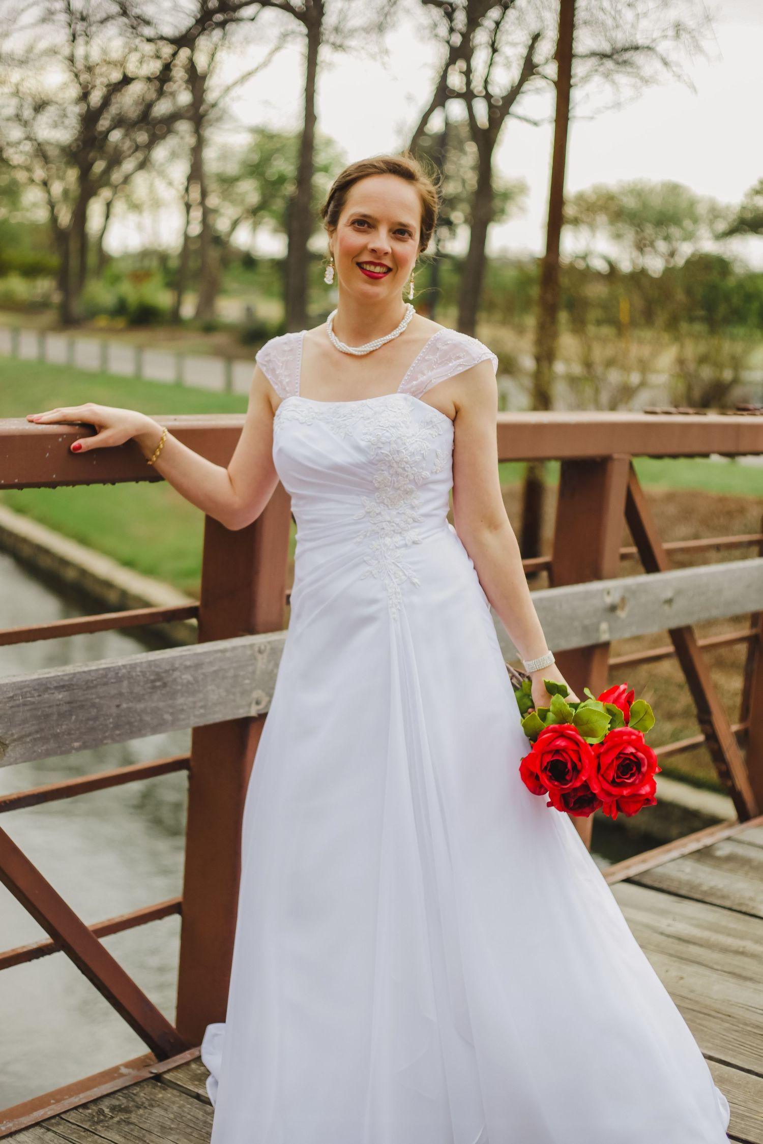 White wedding gown with red roses on rustic wooden bridge.