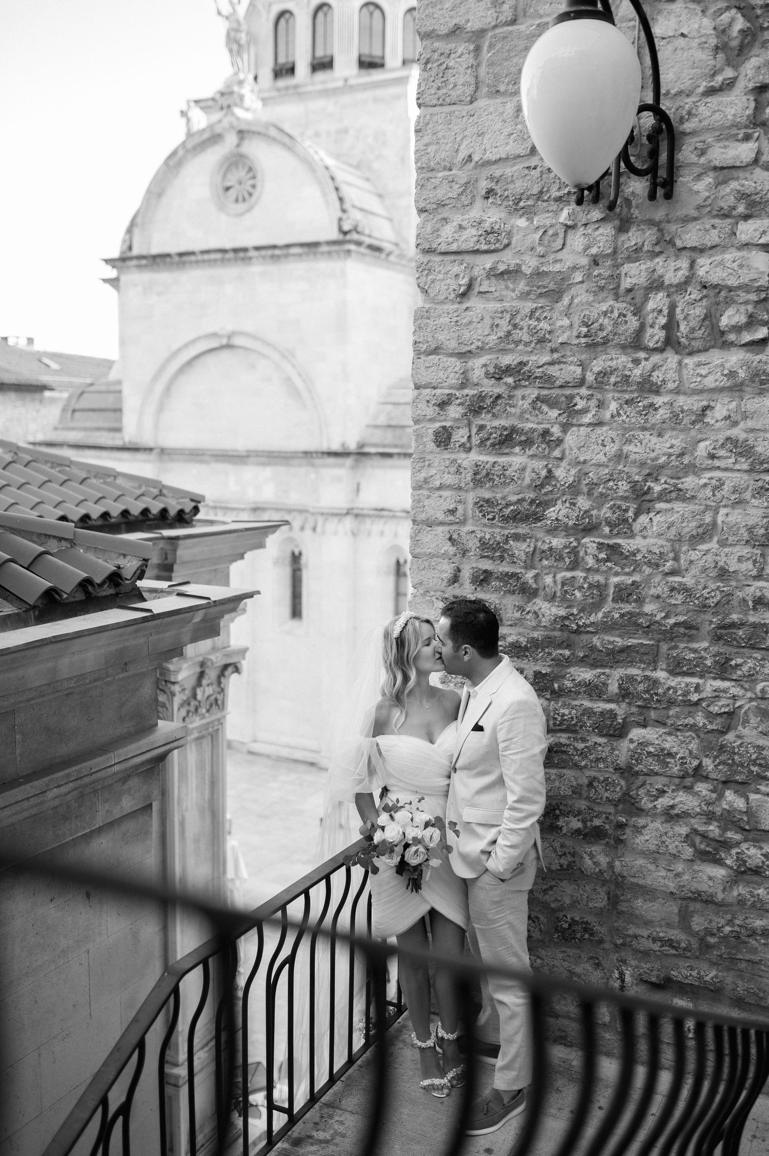 Black and white photo of couple embracing on a Mediterranean balcony with stone walls and church architecture in background.