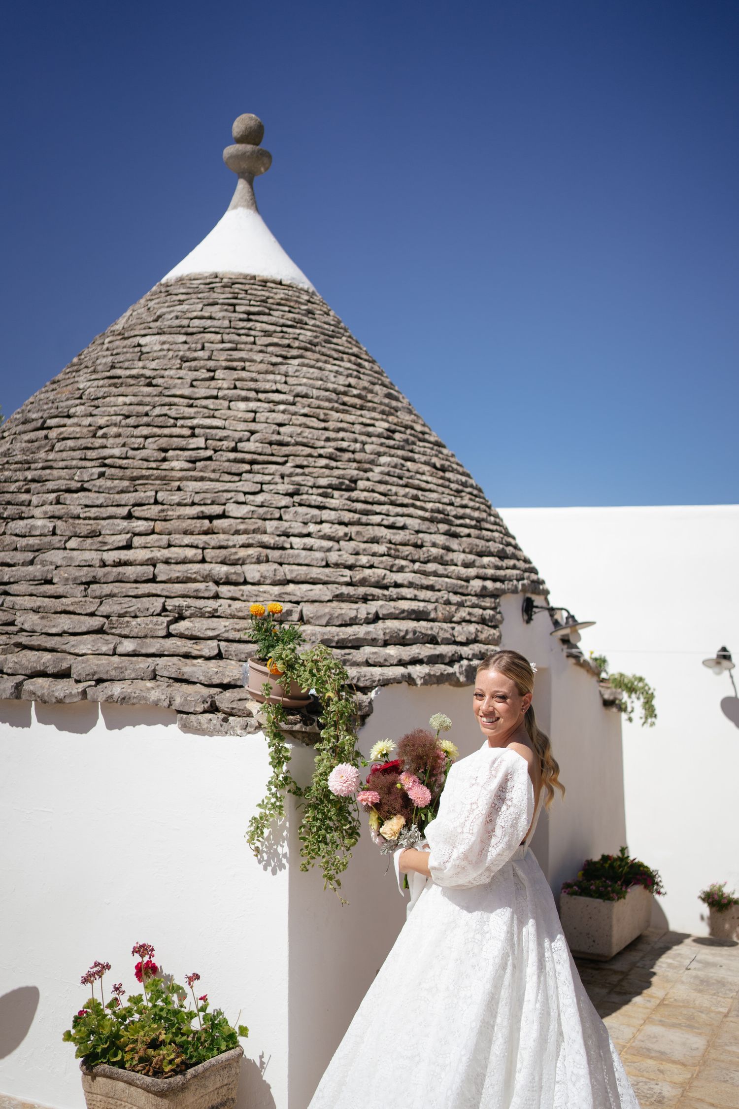Traditional white trulli buildings with stone roofs and flowering plants in Mediterranean setting.